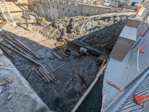 Baustelle mit einem Erdwall, Kränen, Holzpfählen, einem Maschendrahtzaun auf der rechten Seite und Hintergrundelementen wie Gebäuden, Fahrzeugen, Pfählen, Bäumen und Himmel.