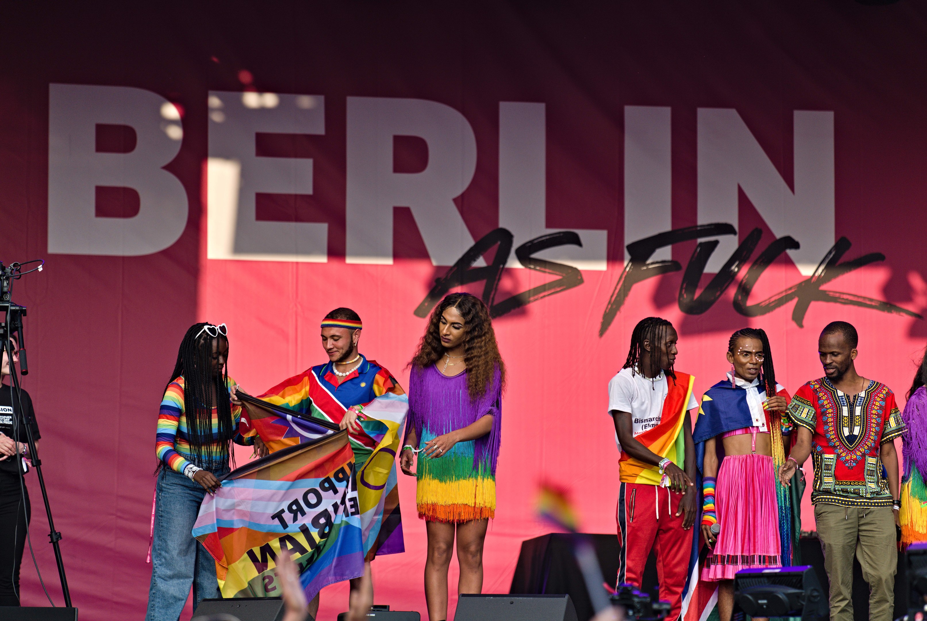 Eine Gruppe von Menschen auf einer Bühne mit einer Frau, die in der Mitte eine Flagge hält, einer Person, die links eine Kamera auf einem Stativ bedient, und einem Banner im Hintergrund mit der Aufschrift 'Berlin als Fuck 2019'.