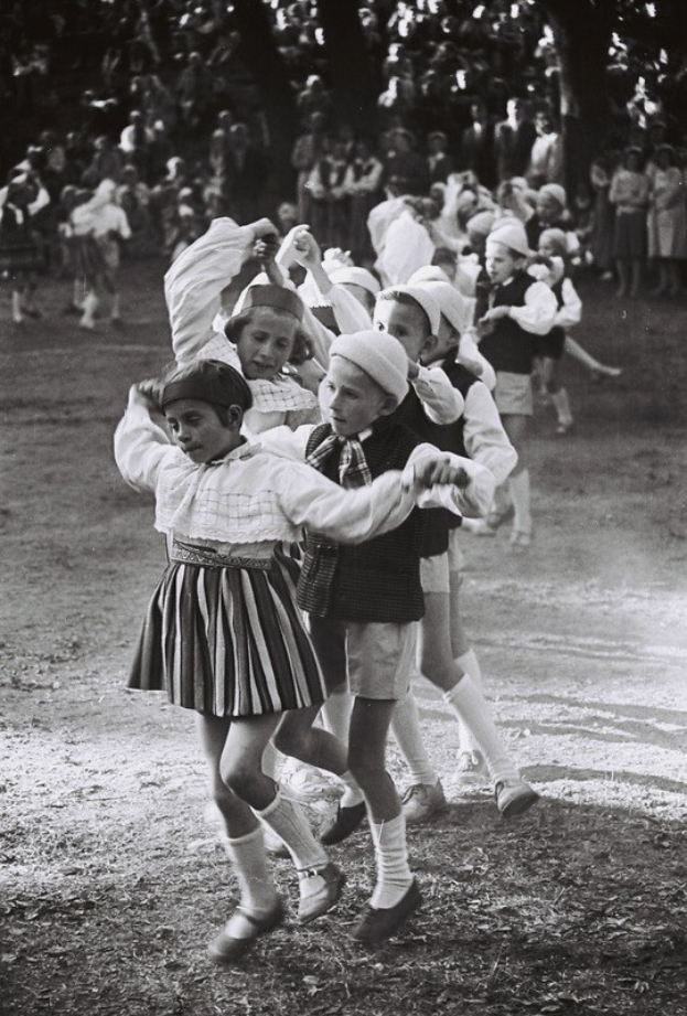 Schwarß-weißes Foto von Kindern, die in einem Park mit Bäumen tanzen und einige stehende Beobachter im Hintergrund.