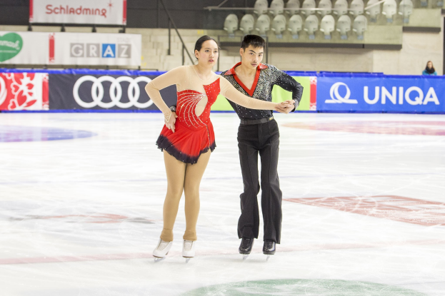 Zwei Eiskunstläufer, ein Mann und eine Frau, gleiten Hand in Hand über eine Eisfläche, mit Zuschauern und Equipment im Hintergrund.