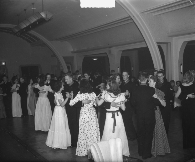 Schwarzes und weißes Bild einer Gruppe von Menschen, die in einem Ballsaal tanzen, mit Deckenbeleuchtung und umgeben von Stühlen.