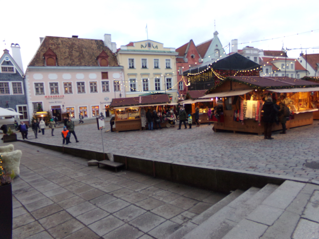 Ein geschäftiger Weihnachtsmarkt in Tallinn, Estland, mit Menschen um dekorierte Stände, festlicher Beleuchtung, Topfpflanzen, Treppen und Gebäuden mit Fenstern unter einem bewölkten Himmel.