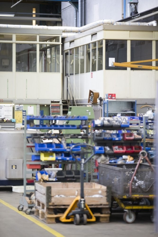 Ein großer Lagerraum voller verschiedener Geräte wie Wagen, Schränke und eine Kiste, mit einer Wand aus Glasfenstern und Rohren im Hintergrund.