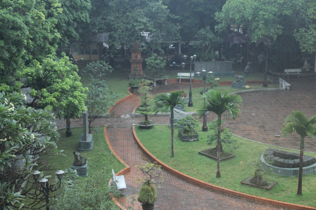 Ein grüner Park mit Bäumen, Pflanzen, Gras, Laternen, Bänken und Skulpturen mit einem Gebäude im Hintergrund.