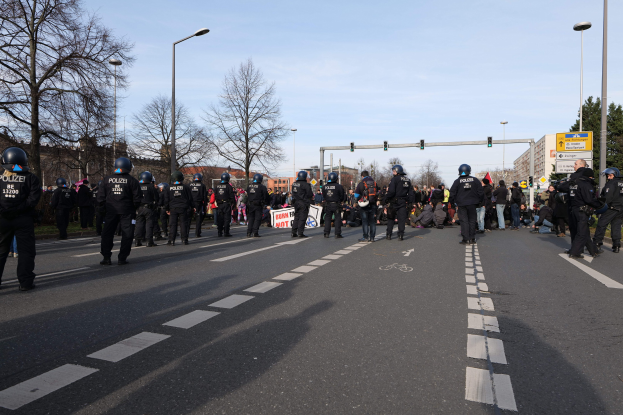 Gruppe von Polizisten in schwarzen Uniformen und Helmen, die an der Straßenseite mit Laternenmasten, Verkehrsampeln, Bäumen, Gebäuden und einem klaren blauen Himmel im Hintergrund stehen.