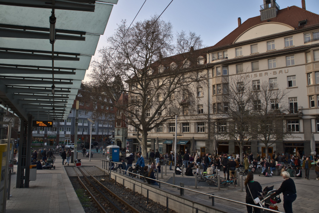 Eine belebte Stadtstraße mit einem Bahnhof auf der linken Seite, umgeben von Gebäuden, Menschen, die gehen, sitzen oder stehen, Bäume, Laternenpfähle und Texttafeln, unter einem sichtbaren Himmel.
