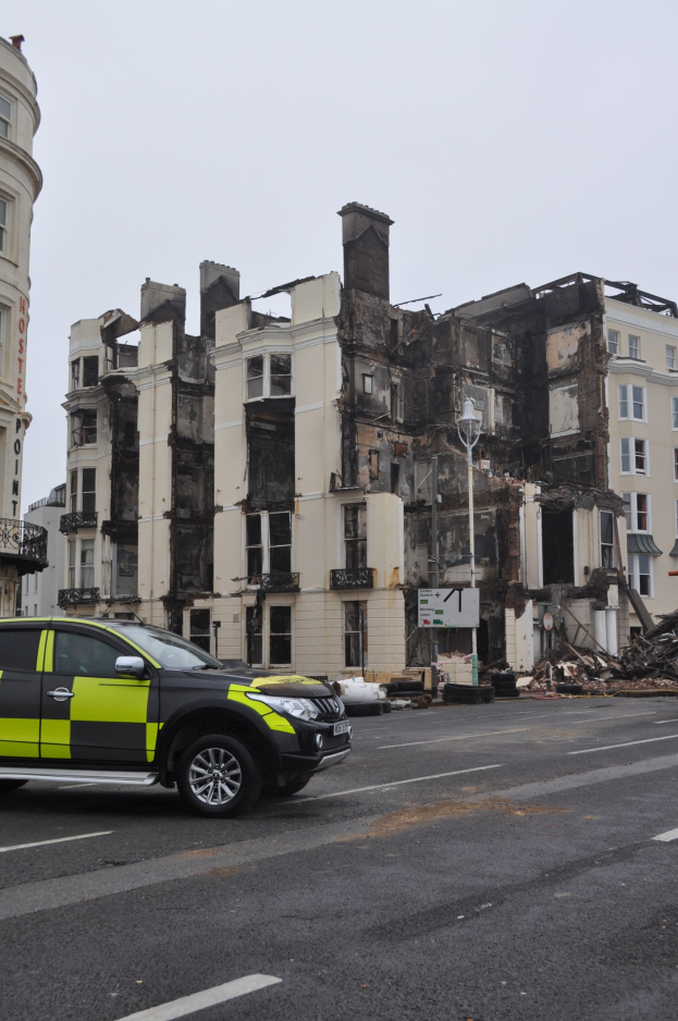 Ein Polizeiwagen vor einem stark beschädigten Gebäude mit zersplitterten Fenstern und verstreuter Trümmer, unter einem sichtbaren Himmel mit benachbarten Gebäuden.