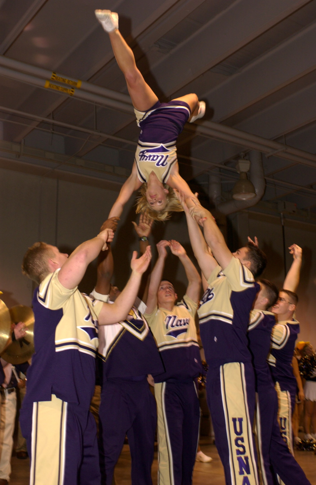Eine Gruppe von Cheerleadern, die einen Stunt in einer Turnhalle ausführen, mit einer Frau in einer Handstandposition in der Mitte und anderen, die um sie herum stehen, einige halten dabei Musikinstrumente auf der linken Seite, und eine Wand mit Rohren und Deckenleuchten im Hintergrund.