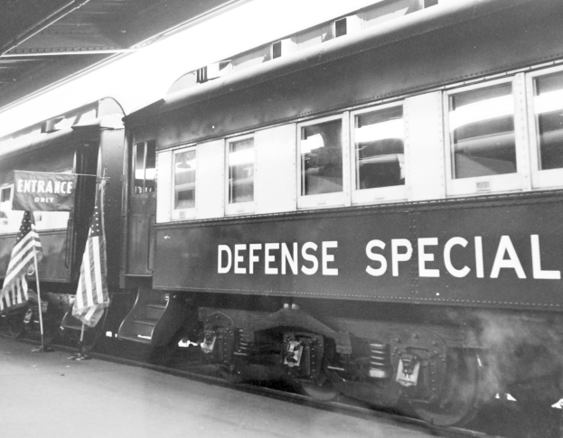 Schwarzes und weißes Foto eines Zuges mit der Aufschrift "Defense Special" auf einem Bahngleis mit zwei Fahnen links und einer Decke oben.