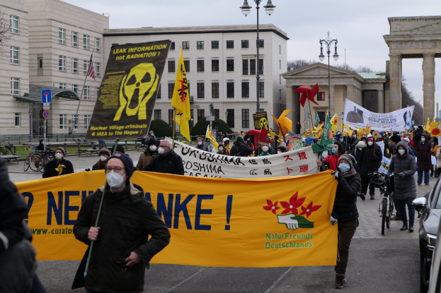 Große Menschenmenge marschiert mit Transparenten und Fahnen auf einer Straße bei einer Demonstration gegen Atomkraft in Deutschland, mit Fahrzeugen, Gebäuden, Bäumen und einem Tor im Hintergrund.