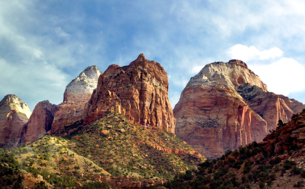 Eine Panoramansicht des Zion-Nationalparks in Utah mit majestätischen Bergen, üppigen Bäumen, steinigem Gelände und einem Himmel mit weißen, flauschigen Wolken.
