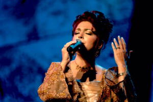 Eine Frau in einem goldenen Kleid singt auf der Bühne der Eurovision Song Contest in ein Mikrofon, mit einem blauen und schwarzen Hintergrund.