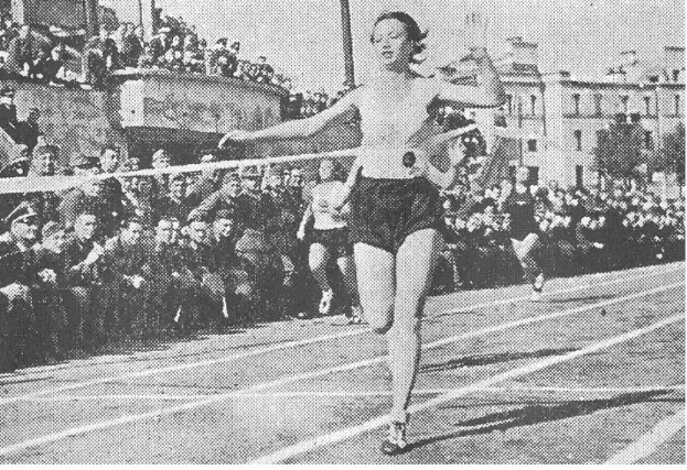 Schwarzes und weißes Foto einer Frau, die auf einer Laufbahn während des Frauen-100m-Finals bei den Olympischen Spielen 1956 läuft, mit Zuschauern, Gebäuden und Pfosten im Hintergrund.