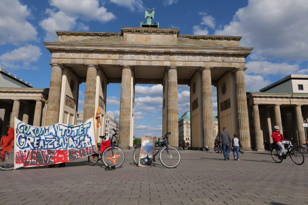 Gruppe von Menschen, die Fahrräder vor dem Brandenburger Tor fahren, einem prächtigen Bogen mit Säulen und einer Statue oben drauf, mit einem Banner und Gebäuden im Hintergrund.