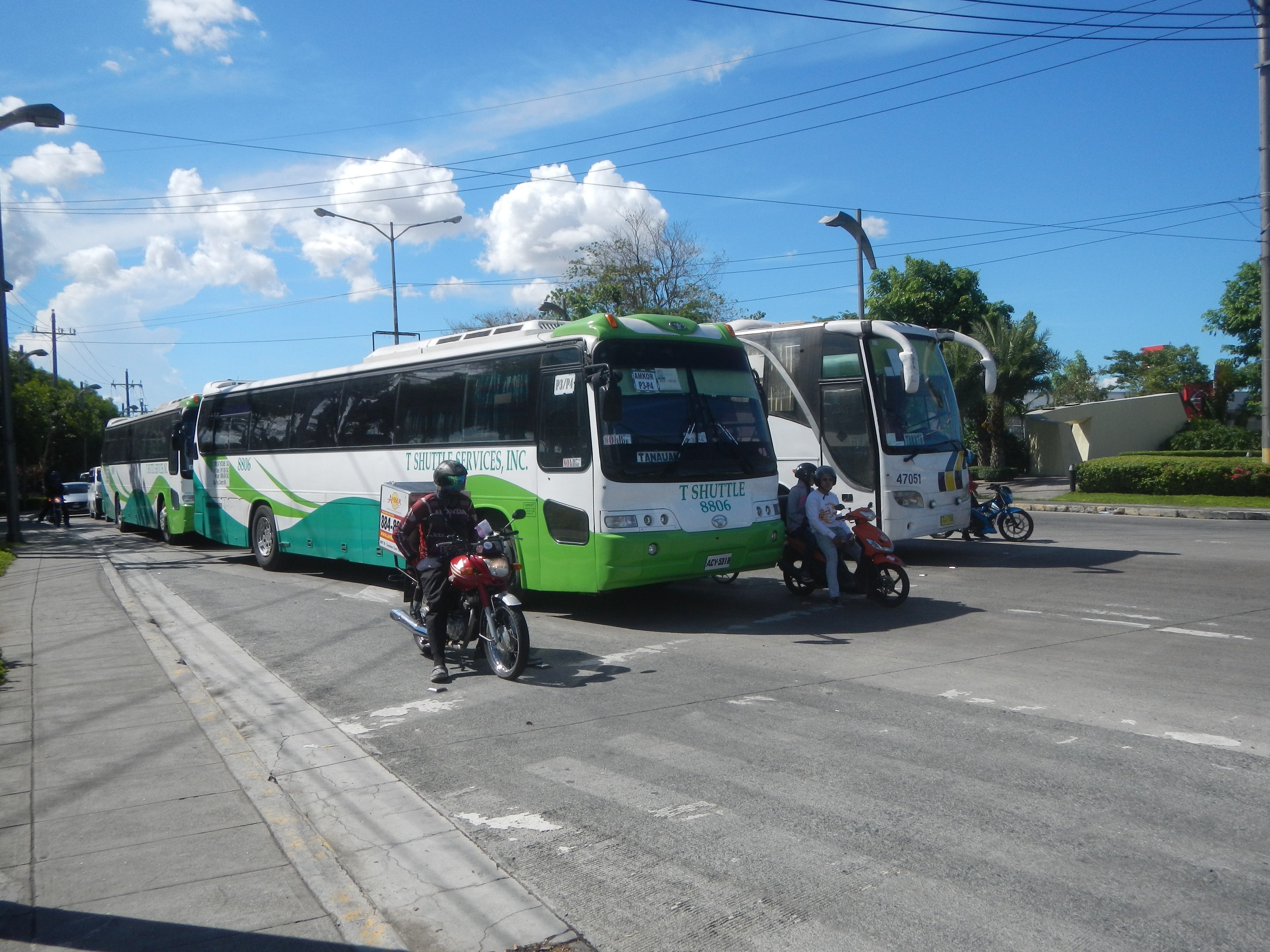 Ein grüner und weißer Shuttlebus steht am Straßenrand mit Motorrädern davor, ein grasbewachsener Fußweg links daneben und Gebäude, Bäume und Laternenmasten im Hintergrund unter einem klaren blauen Himmel.