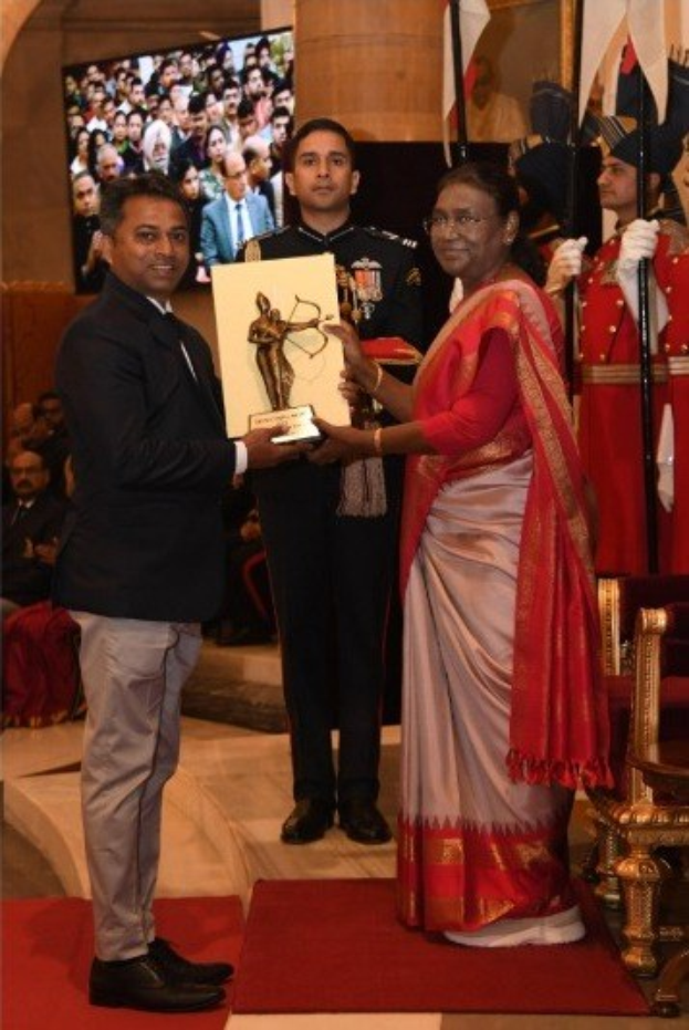 Ein Mann in einem schwarzen Blazer, weißem Hemd und grauen Hosen hält eine Trophäe, während er neben einer Frau in einem ashesfarbenen Sari auf einem roten Teppich an einer Preisverleihung steht, mit Menschen, Stühlen, Fahnen und einem Bildschirm im Hintergrund.