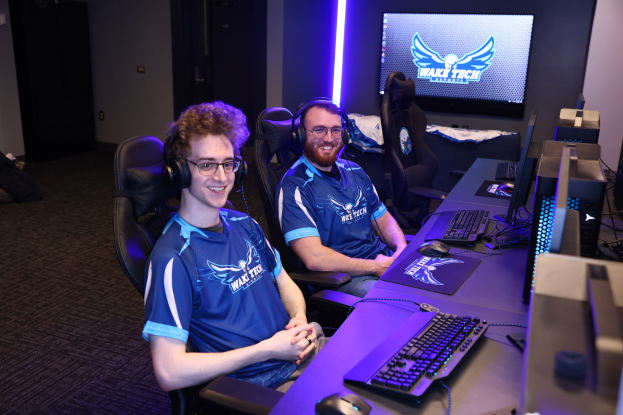 Zwei Männer mit Headsets lächeln, während sie an einem Schreibtisch mit Computermonitoren, Tastaturen und anderem Equipment sitzen und ein Spiel in einem Raum mit einem Bildschirm und Lichtern im Hintergrund spielen.