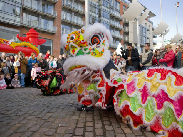 Ein farbenfrohes chinesisches Neujahrsfest in Amsterdam mit einem Löwen Tanz und einer Menge Schaulustiger, einige machen Fotos, vor einem Hintergrund aus Gebäuden, Laternenmasten und einem klaren blauen Himmel.