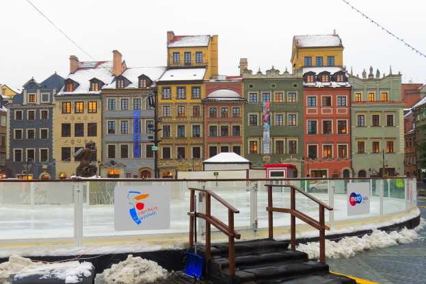 Eislaufbahn in einem Stadtplatz umgeben von Geb├Ąuden, Treppen, Gel├Ąndern, Informationsschildern, schneebedeckter Erde, Laternen, B├Ąumen und einem Himmelhintergrund.