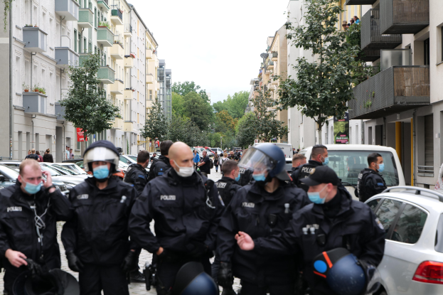 Polizisten in Masken und Helmen gehen eine Straße mit parkenden Autos entlang, flankiert von Gebäuden mit Balkonen und Geländern, unter einem klaren Himmel mit Bäumen im Hintergrund.