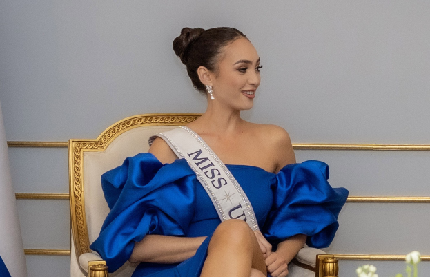 Frau in einem blauen Kleid und einem "Miss Universe 2019"-Band, die auf einem Stuhl sitzt und lächelt, umgeben von Blumen und einer Wand im Hintergrund.