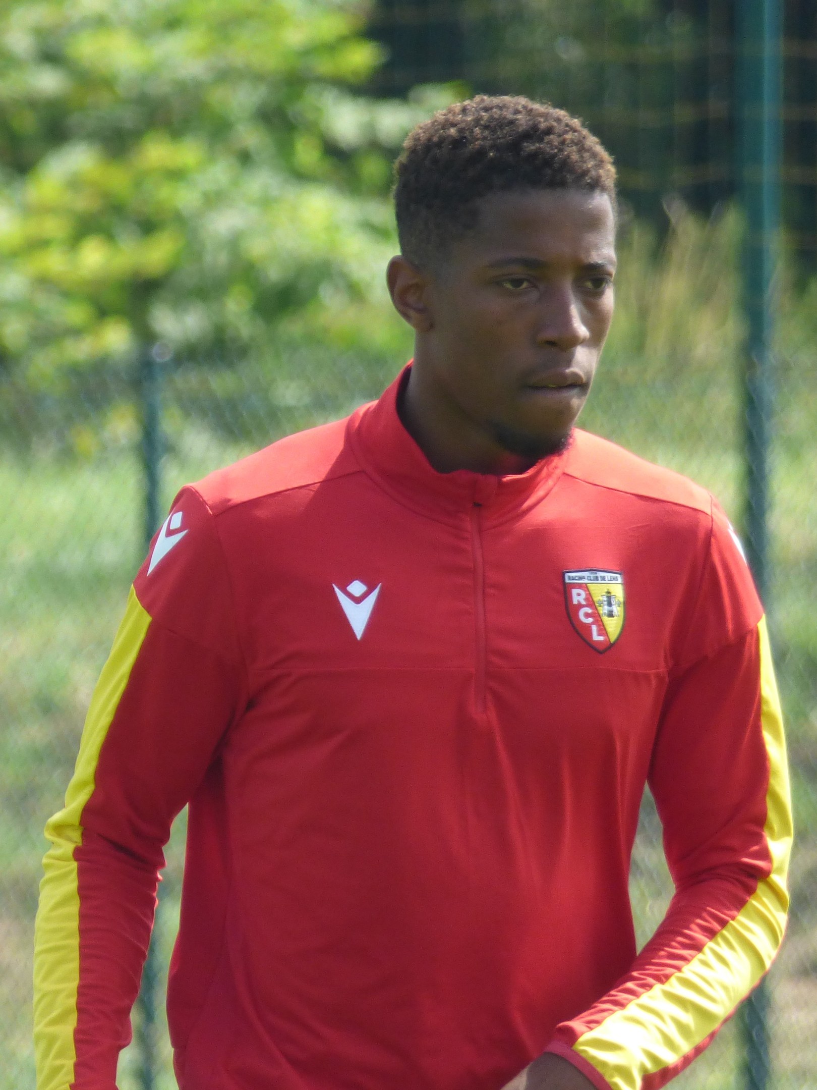 Ein junger Mann in einem roten und gelben Fußballtrikot steht auf einem Feld, hält sich entschlossen an einem Zaun fest, Bäume sind im Hintergrund zu sehen.