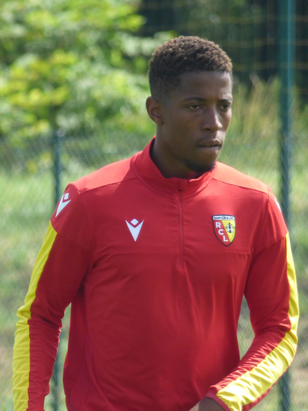 Ein junger Mann in einem roten und gelben Fußballtrikot steht auf einem Feld, hält sich entschlossen an einem Zaun fest, Bäume sind im Hintergrund zu sehen.