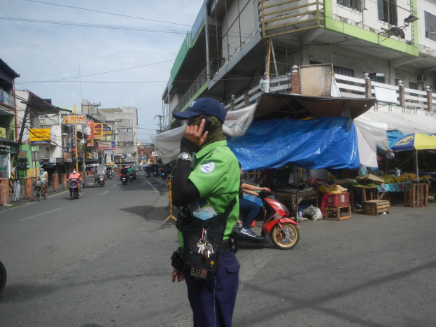Ein Polizist in grüner und blauer Uniform mit blauer Mütze steht an der Seite einer Straße und spricht auf einem Handy, während Fahrzeuge auf der Straße hinter ihm fahren und Stände zu seiner Rechten verschiedene Gegenstände ausstellen.