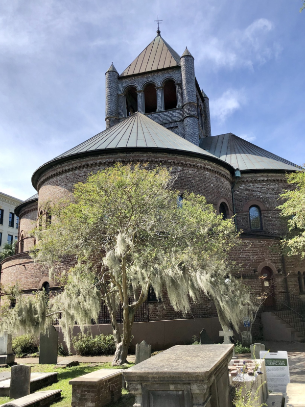 Außenansicht der Kirche des Heiligen Grabes in Charleston, South Carolina, mit einem Turm, einem Baum, umgeben von Gebäuden, einer Treppe mit Geländer, Gras und einer Texttafel, unter einem bewölkten Himmel.