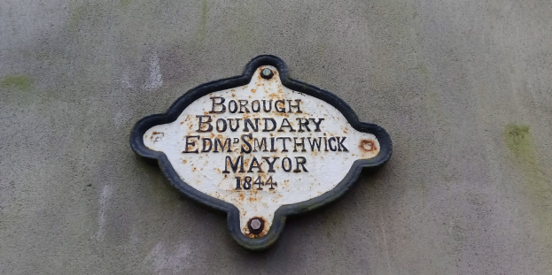 Schwarze Metalltafel mit weißer, fetter, zentrierter Schrift, die "Gemeindegrenze Edm Smithwick Bürgermeister" auf der Seite eines Gebäudes liest.