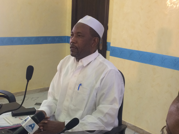 Ein Mann in einem weißen Kleid und Hut sitzt an einem Tisch mit Mikrofonen, spricht, während Papiere und Gegenstände auf dem Tisch zu sehen sind, mit einem anderen Mann mit Brille zu seiner Rechten, in einem Raum mit einer Wand und einer Tür im Hintergrund.