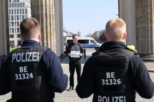Drei Polizeibeamte stehen vor einem Gebäude, ein Mann hält ein Papier in der Hand und ist von Säulen und Fahrzeugen, Menschen, Bäumen und Gebäuden umgeben.
