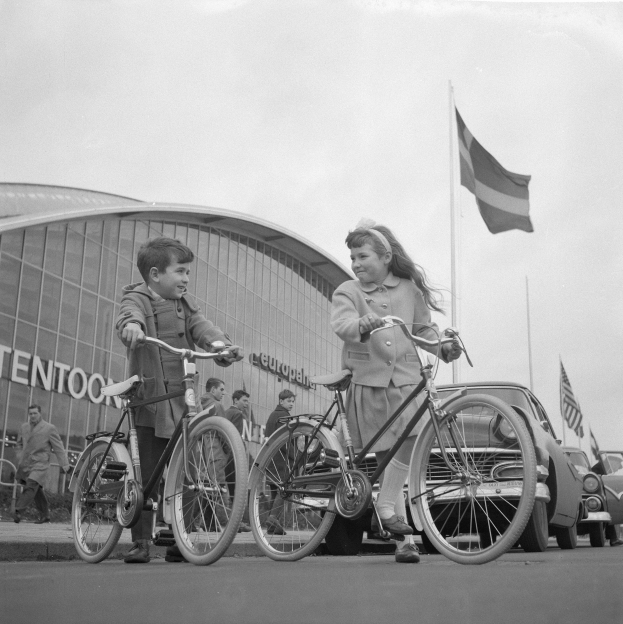 Schwarz-weiß-Foto von zwei Kindern, die vor einem Gebäude Fahrräder fahren, mit einigen Menschen, Fahrzeugen und Flaggen im Hintergrund.