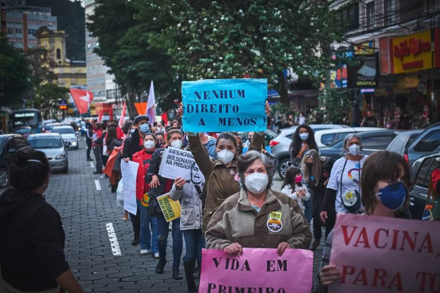 Eine Gruppe von Menschen mit Masken und Schildern protestiert auf einer Straße, umgeben von Fahrzeugen, Bäumen, Gebäuden und Texttafeln, mit dem Himmel im Hintergrund.