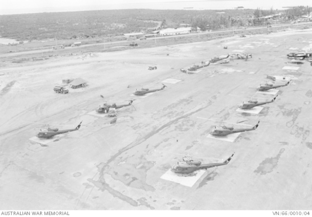 Schwarzes und weißes Foto mehrerer Militärhubschrauber auf dem Boden mit umliegenden Fahrzeugen, Schuppen, Polen, Bäumen und Wasser sowie Text unten.