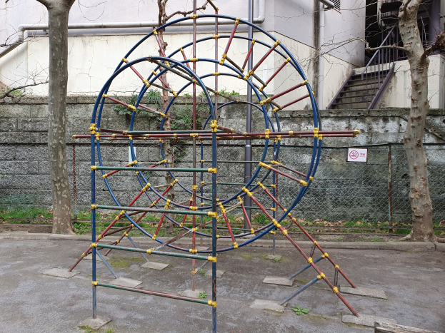 Spielplatz mit einem großen Metall-Klettergerüst, umgeben von Bäumen und einer Maschendrahtzaun, mit Treppen mit Geländern, die zu einem Gebäude mit Fenstern und Rohren führen, einem Schild an einem Pfahl und grasbewachsenem Boden.