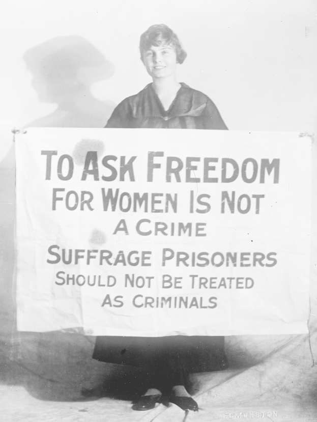 Schwarz-weißes Foto einer Frau, die ein Banner hält, auf dem steht: "Für die Freiheit der Frauen zu kämpfen ist kein Verbrechen, Suffragetten sollten nicht wie Kriminelle behandelt werden." und dabei lächelt.
