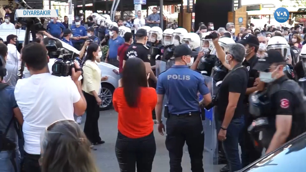 Eine Gruppe von Menschen vor einer Reihe von Polizisten in Helmen und mit Kameras, mit Fahrzeugen, Gebäuden und Schildern im Hintergrund, die 'türkische Polizei im Konflikt mit Demonstranten in Ankara' lesen.