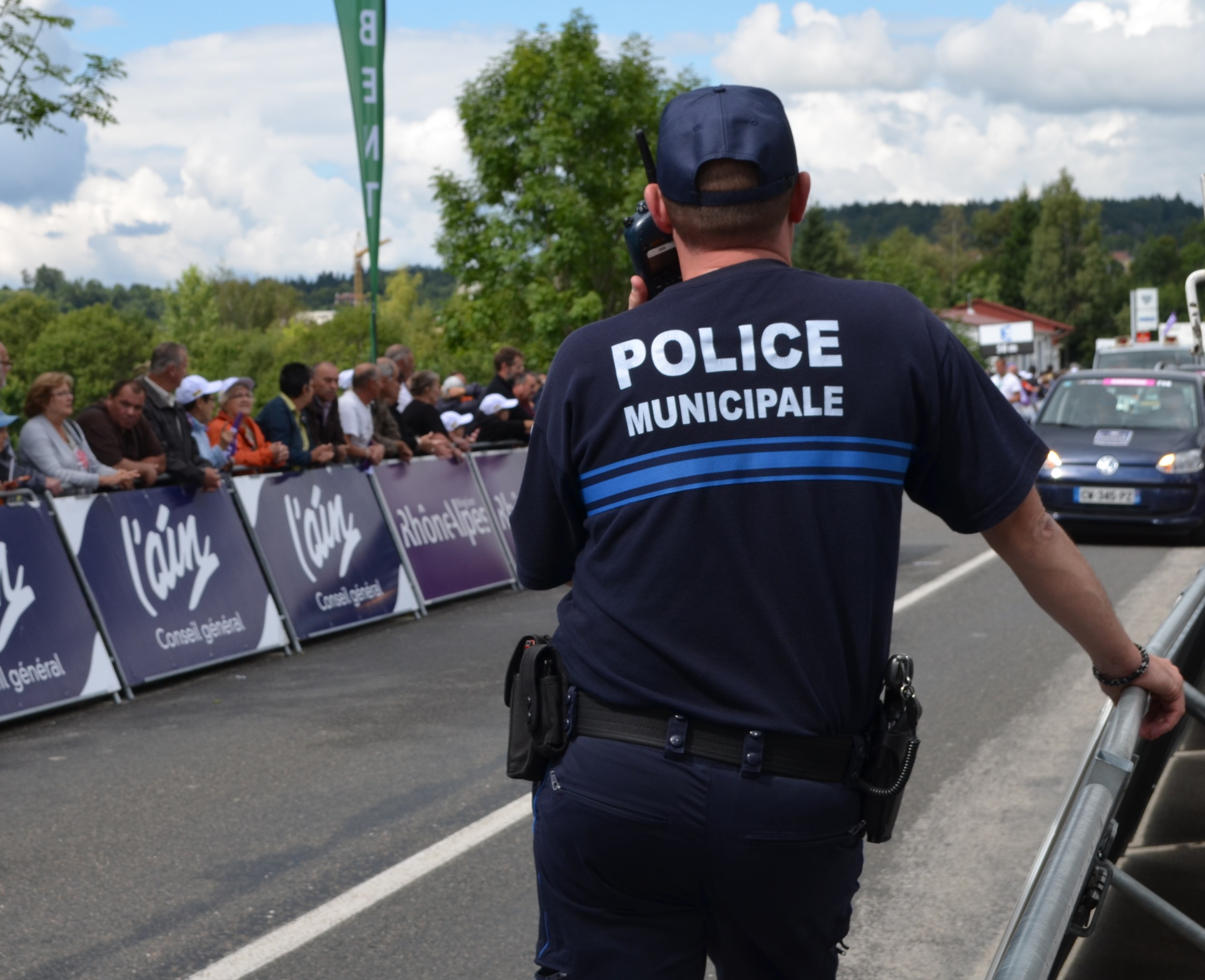 Polizist in Uniform mit Mütze und Funkgerät vor einer Menge, Schildern, Fahrzeugen, Bäumen, Gebäuden und bewölktem Himmel.