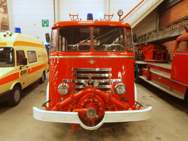 Zwei Feuerwehrautos nebeneinander in einer Garage geparkt mit einem Rettungswagen links, einer Treppe an der rechten Wand und Schildern und Rollläden im Hintergrund.