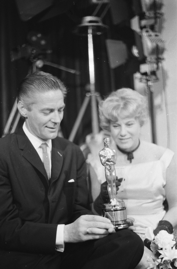 Ein Mann in einem schwarzen Blazer, weißem Hemd und Krawatte hält eine Oscar-Statue in der Hand und sitzt neben einer Frau, die einen Blumenstrauß hält, beide in formeller Kleidung, mit ein paar Lichtern im Hintergrund, in Schwarz-Weiß.