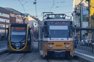Zwei gelbe und weiße Straßenbahnen auf einer Stadtstraße mit hohen Gebäuden, Fahrzeugen, Geländern, Polen, Schildern, Bäumen, einem Hügel und dem Himmel im Hintergrund.