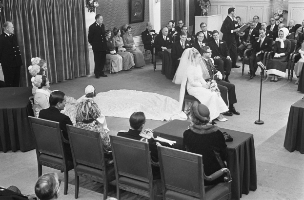 Schwarzes und weißes Foto einer Hochzeitszeremonie mit sitzenden und stehenden Gästen, einem Mann, der in ein Mikrofon spricht, einer Frau mit einem Blumenstrauß, einem Podium mit einem Mikrofon auf der rechten Seite, Blumenschmuck und einem Vorhang auf der linken Seite, einem gerahmten Foto und einer Tür im Hintergrund.