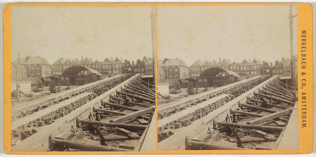 Ein altes Schwarz-Weiß-Foto einer Bahnschiene mit Gebäuden im Hintergrund, Text auf der rechten Seite lesend "Merkelbach & Co, Co. Amsterdam" und verstreute hölzerne Gegenstände im Vordergrund.