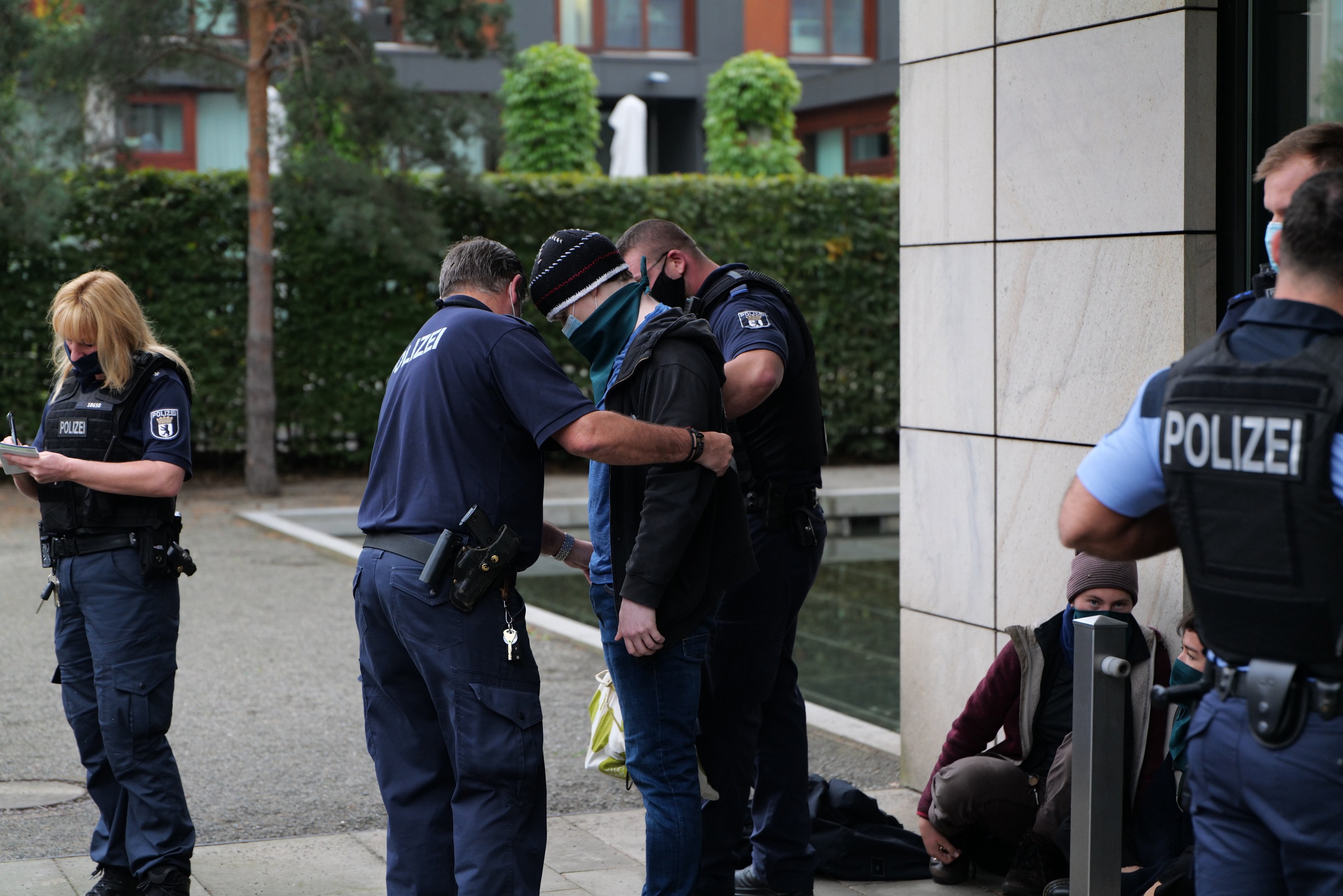 Deutsche Polizeibeamte, die in einer Gruppe vor einem Gebäude stehen, mit einer Frau, die ein Buch und einen Stift hölt, und Menschen in der Nähe, umgeben von Pflanzen und urbanen Strukturen.