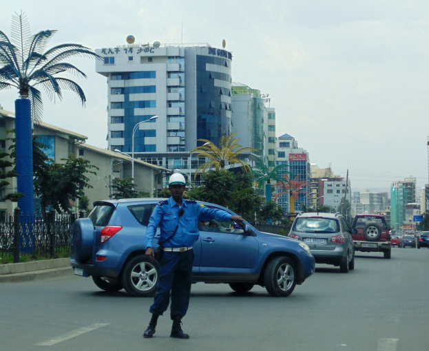Polizist in blauer Uniform und weißem Helm auf einer Stadtstraße mit Fahrzeugen, Gebäuden, Bäumen und einem klaren Himmel im Hintergrund.