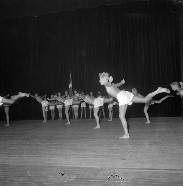 Schwarzes Bild einer Gruppe, die auf einer Bühne Gymnastik macht, mit einem Vorhang und einer Flagge im Hintergrund.