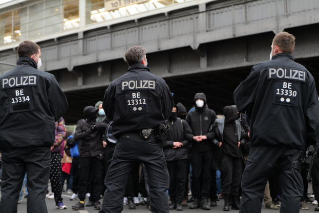 Polizeibeamte in schwarzen Uniformen und Masken vor einer Menge während einer Demonstration, mit einer Stadtbrücke und einem Gebäude im Hintergrund.