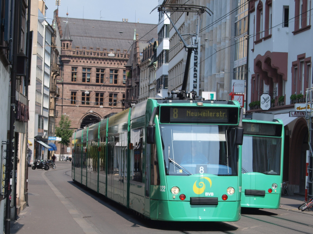Zwei grüne Straßenbahnen fahren eine Stadtstraße entlang, flankiert von hohen Gebäuden, mit parkenden Fahrrädern und Fußgängern auf den Gehwegen, unter einem klaren blauen Himmel mit einem Baum im Hintergrund.