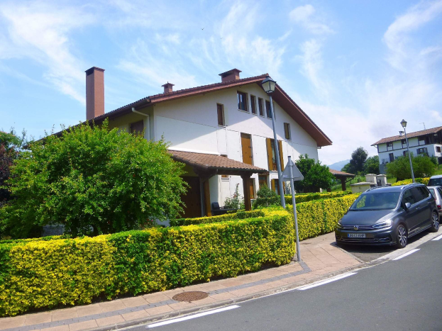Ein zweistöckiges Villa zum Verkauf am Lago di Garda, Italien, mit einem Auto auf der Straße davor, umgeben von Grünflächen und Laternen unter einem teilweise bewölkten Himmel.
